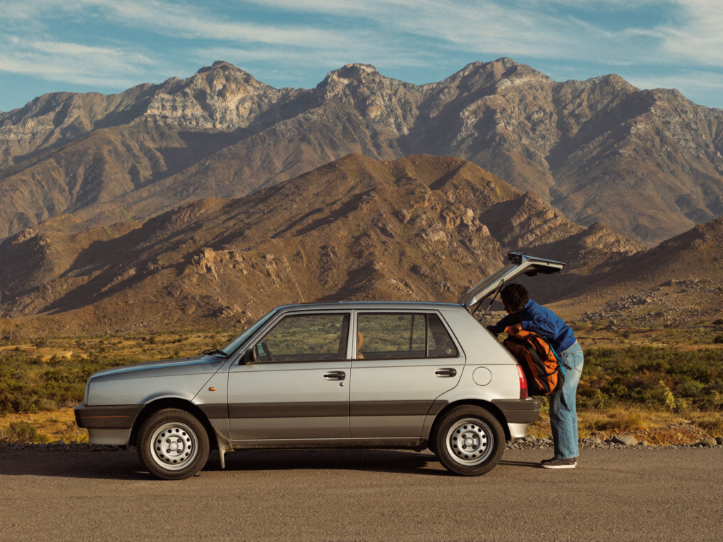 A traveler loading a bag into a rental car parked on a road with Anti-Atlas mountains in the background