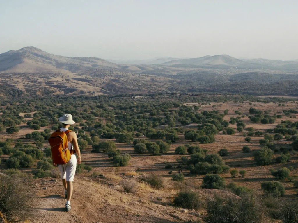 Hiker with an orange backpack on a dry trail overlooking a valley of argan trees in southern Morocco