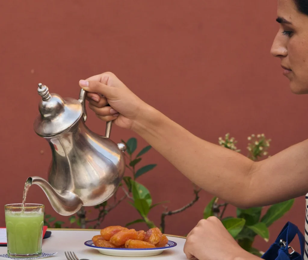 Traveller sitting alone at a cafe table pouring tea from a silver teapot into a glass beside a plate of dates